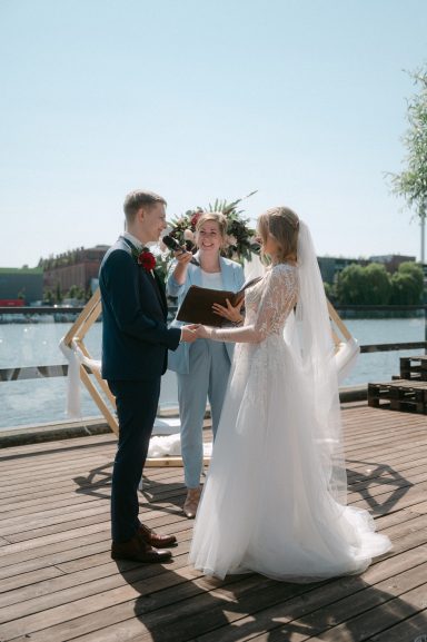 Freie Rednerin traut ein Brautpaar bei sonnigem Wetter auf einem Steg am Wasser in moderner Outdoor-Kulisse. Freie Trauung Berlin.